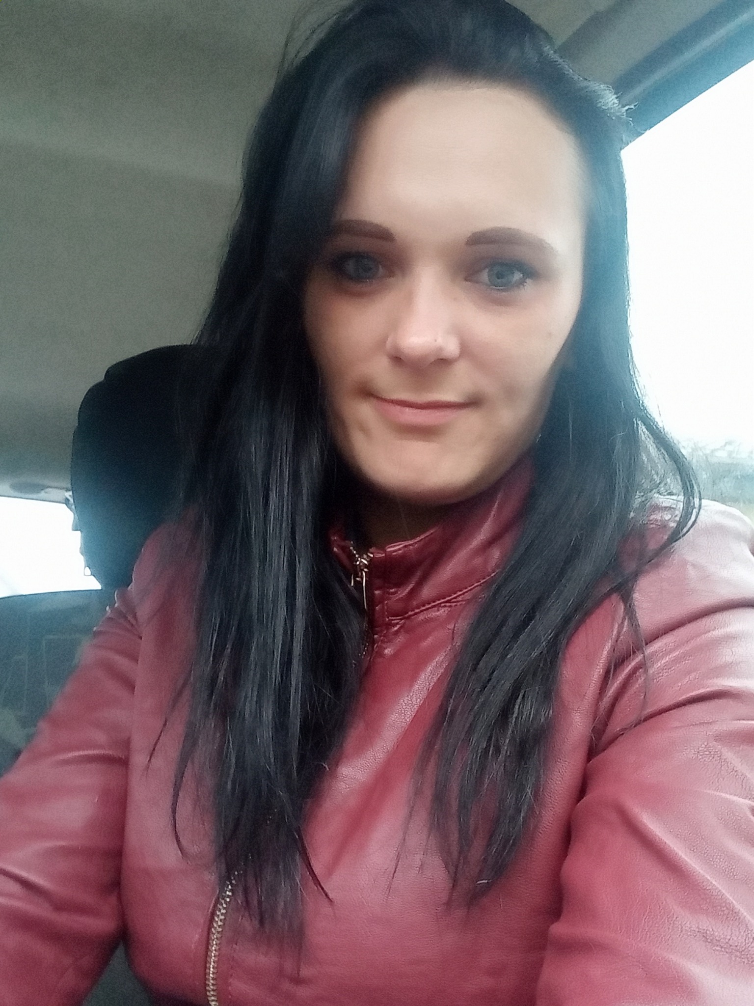 Portret kobiety z długimi, ciemnymi włosami w czerwonej skórzanej kurtce, zrobiony w samochodzie, z widocznym fragmentem tylnego siedzenia.