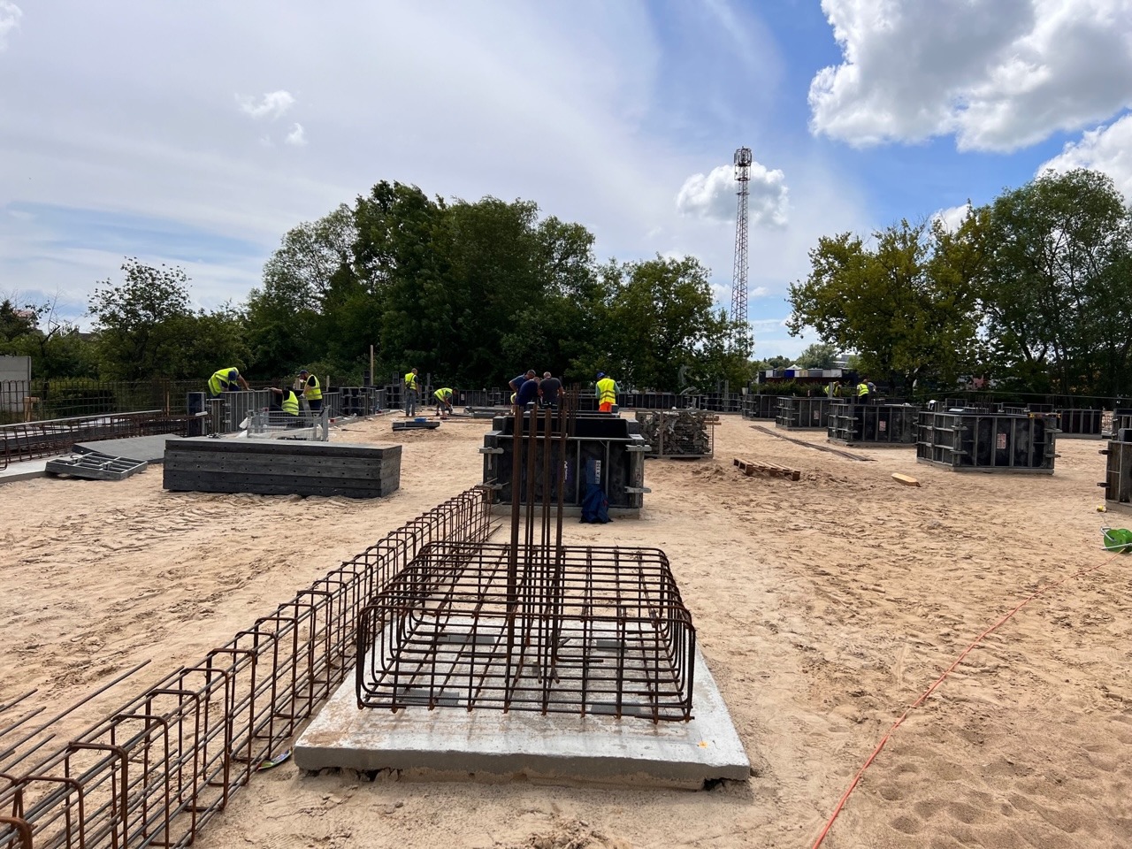 Budowa fundamentów: zbrojenie i szalunki pod słupy na piaszczystym podłożu, pracownicy w kamizelkach odblaskowych w tle, widok na teren budowy w słoneczny dzień.