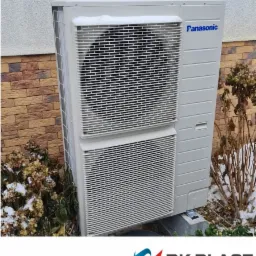 Zewnętrzna jednostka klimatyzacji Panasonic, lekko ośnieżona, zamontowana przy budynku z cegły w Suwałkach. Widoczne krzewy z resztkami śniegu.