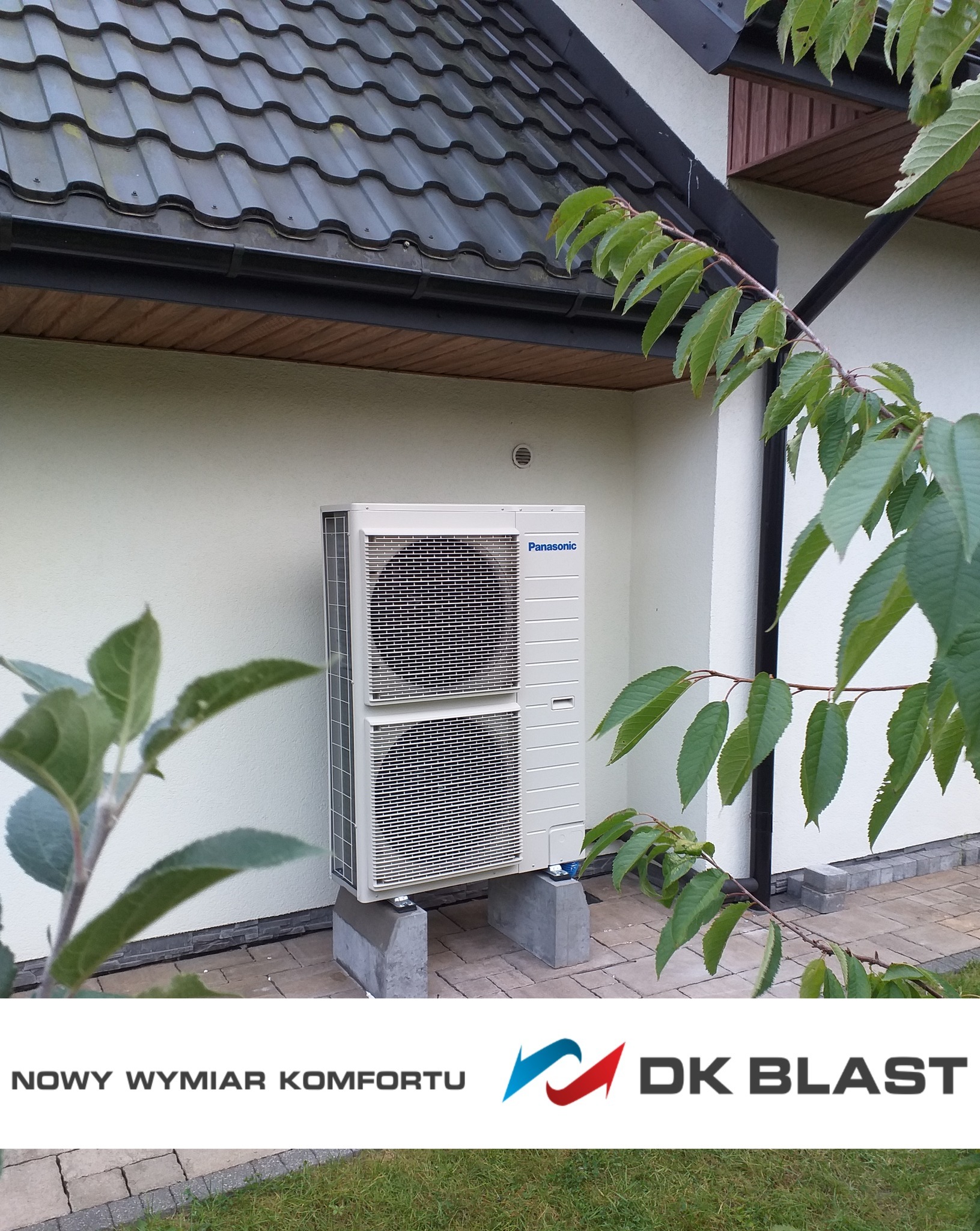 Zewnętrzna jednostka pompy ciepła Panasonic na betonowych podstawach przy ścianie domu, z widoczną gałęzią drzewa z liśćmi na pierwszym planie i logotypem firmy instalacyjnej w dolnej części kadru.