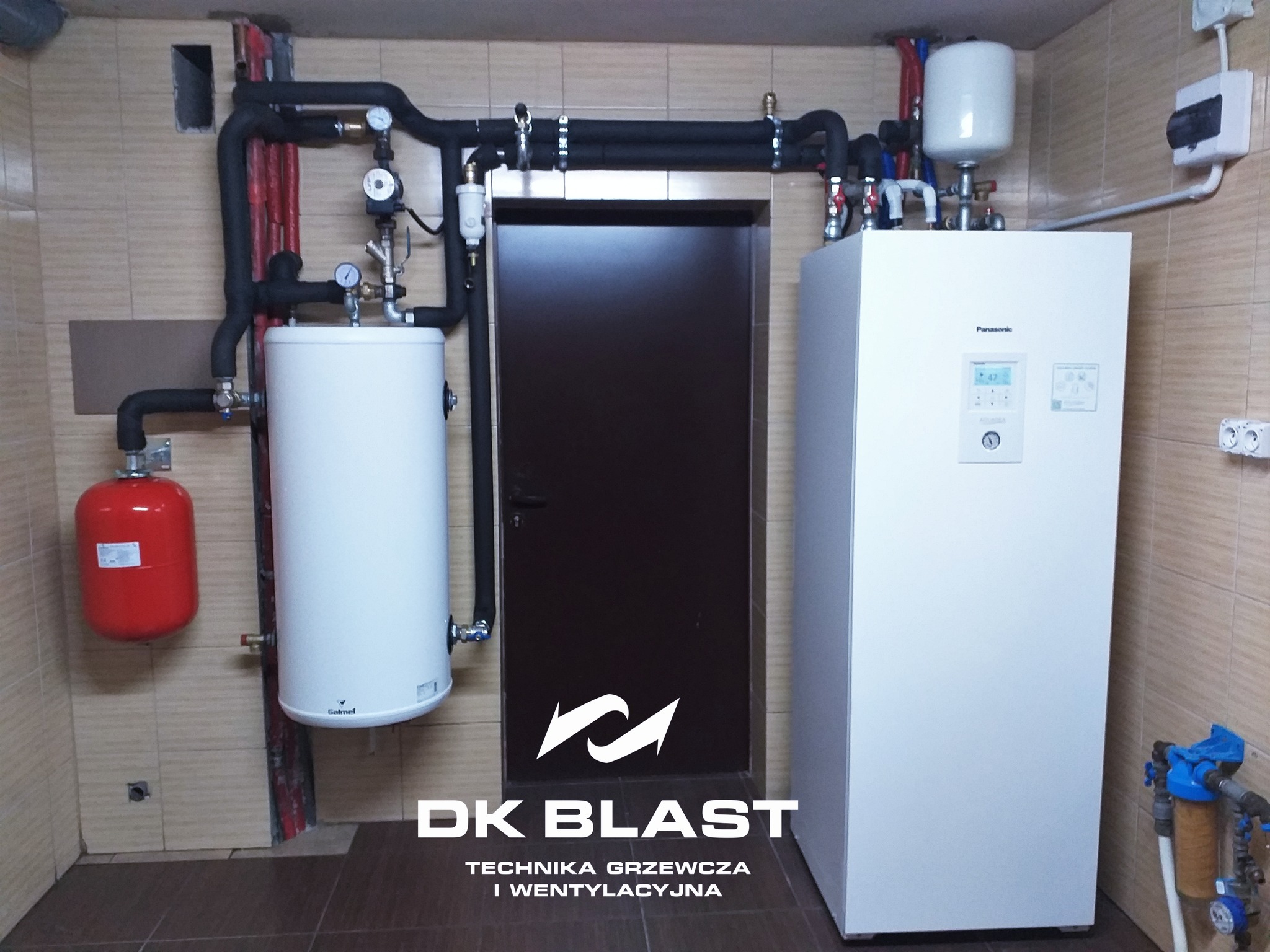 Biały bojler Galmet i pompa ciepła Panasonic z podłączonymi rurami w kotłowni z brązowymi płytkami, widoczny również czerwony zbiornik wyrównawczy, na dole logo DK Blast