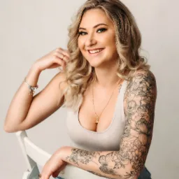 Portret uśmiechniętej blondynki w jasnym topie i dżinsowych szortach, z tatuażami na ramieniu, siedzącej na białym krześle na jasnym tle.