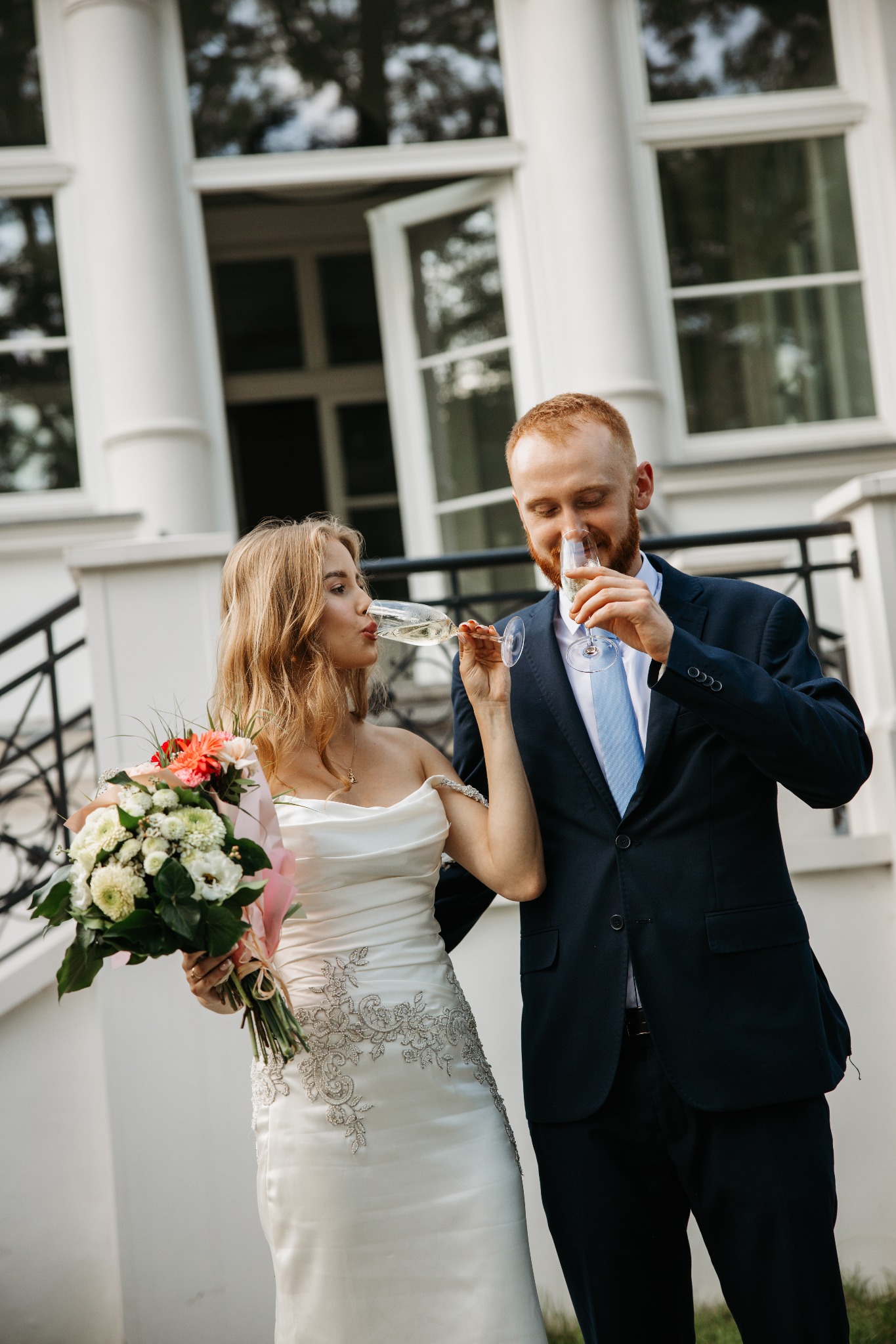 Para młoda w strojach ślubnych pije szampana przed białym budynkiem z otwartymi oknami. Panna młoda trzyma bukiet kwiatów.