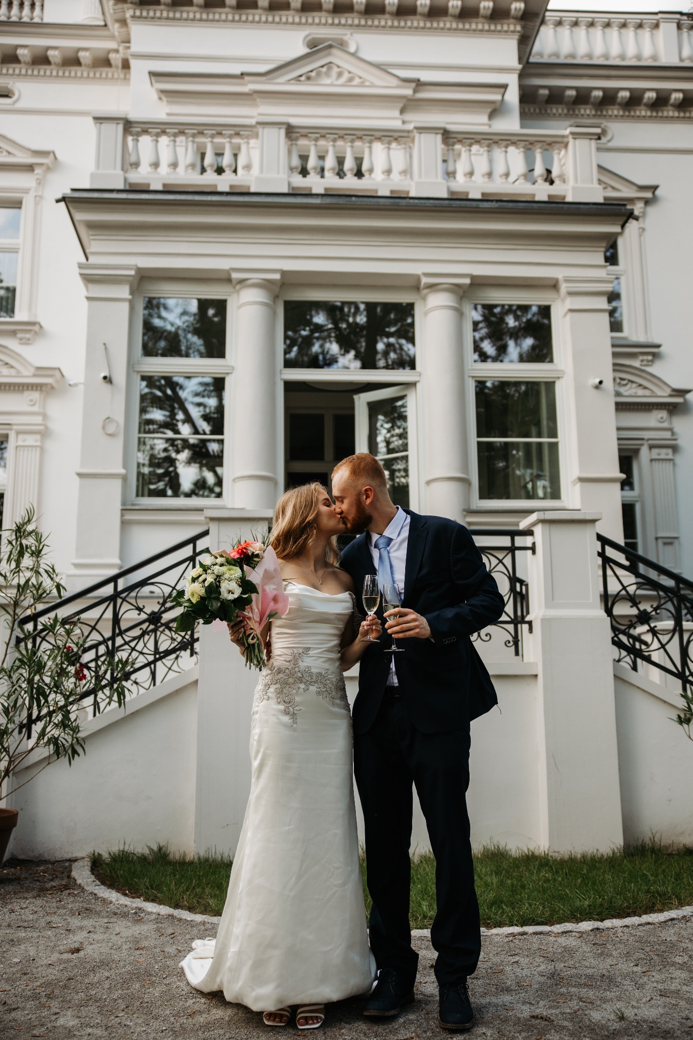 Pocałunek pary młodej z szampanem na tle klasycznej architektury z kolumnami i balkonem.