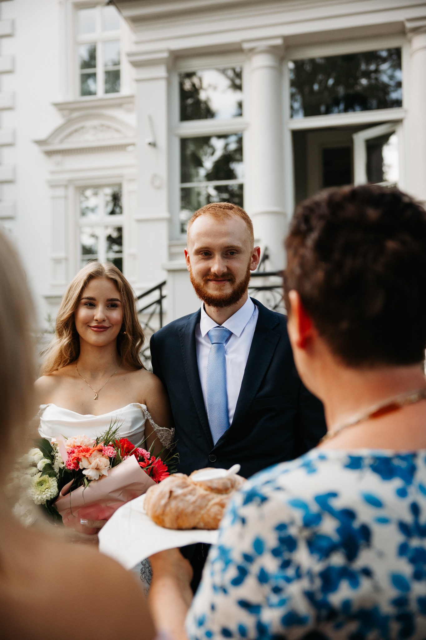 Para młoda w otoczeniu gości, panna młoda trzyma bukiet kwiatów, a gość podaje chleb na powitanie, na tle białego budynku.
