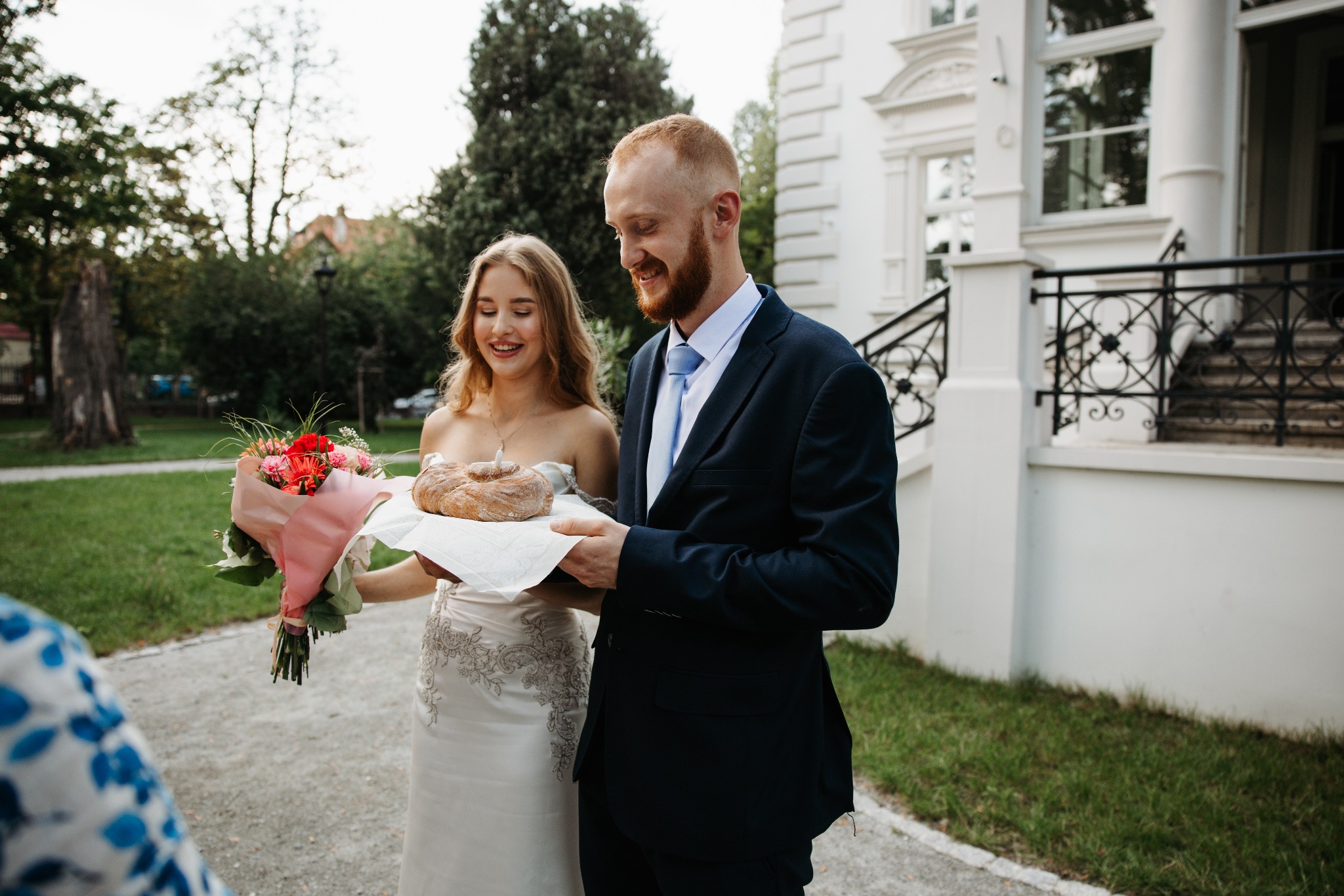Uśmiechnięta para młoda w plenerze: panna młoda w białej sukni trzyma bukiet kwiatów, pan młody w granatowym garniturze podaje chleb weselny na białej serwetce, w tle biały budynek z balustradą...