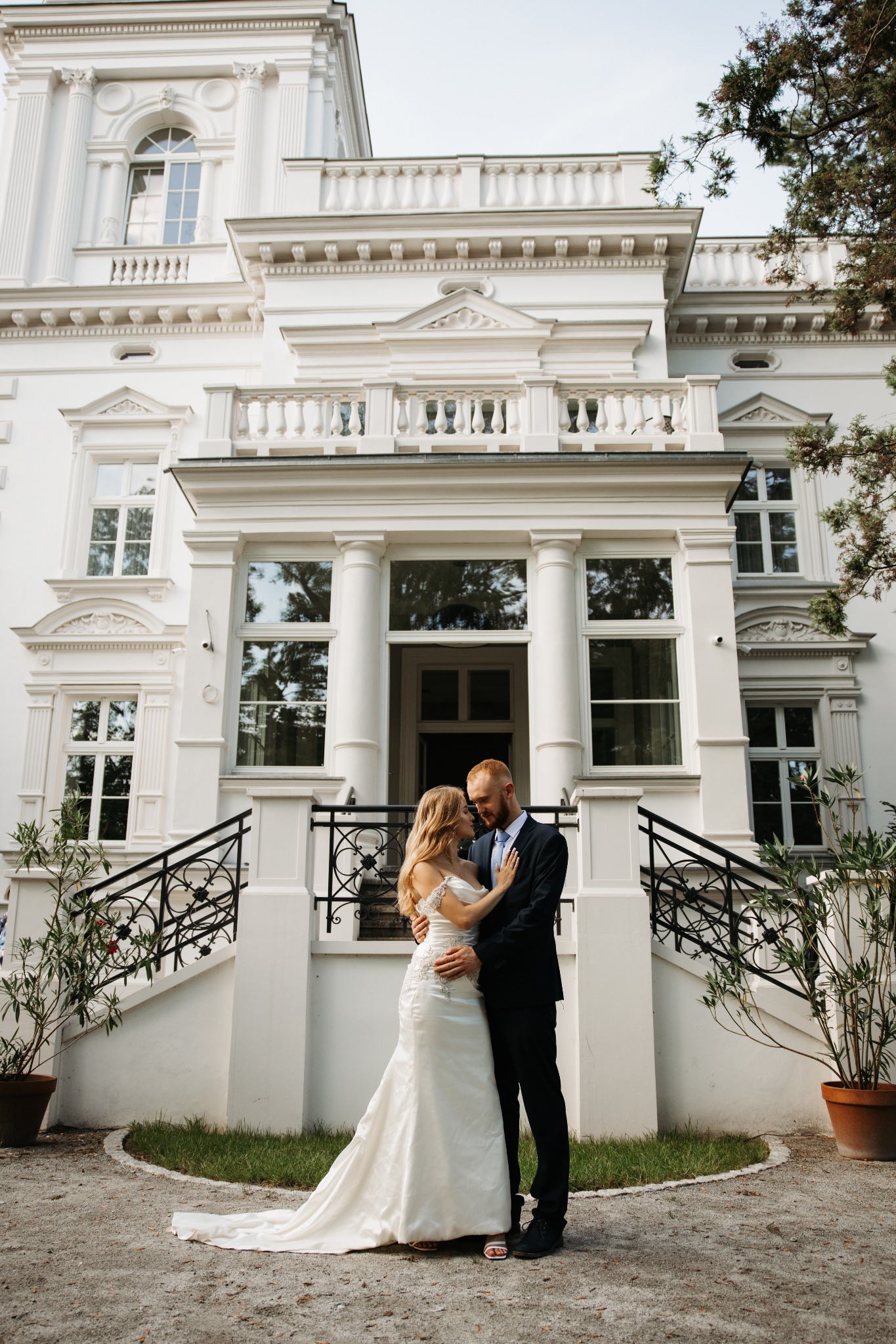 Para młoda obejmująca się na tle białego, klasycystycznego budynku z kolumnami i schodami z kutą balustradą.