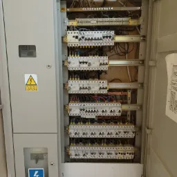 Otwarta skrzynka bezpiecznikowa w szarej obudowie z widocznymi bezpiecznikami, przewodami i naklejką ostrzegawczą o napięciu elektrycznym oraz schematem obwodów po prawej stronie.