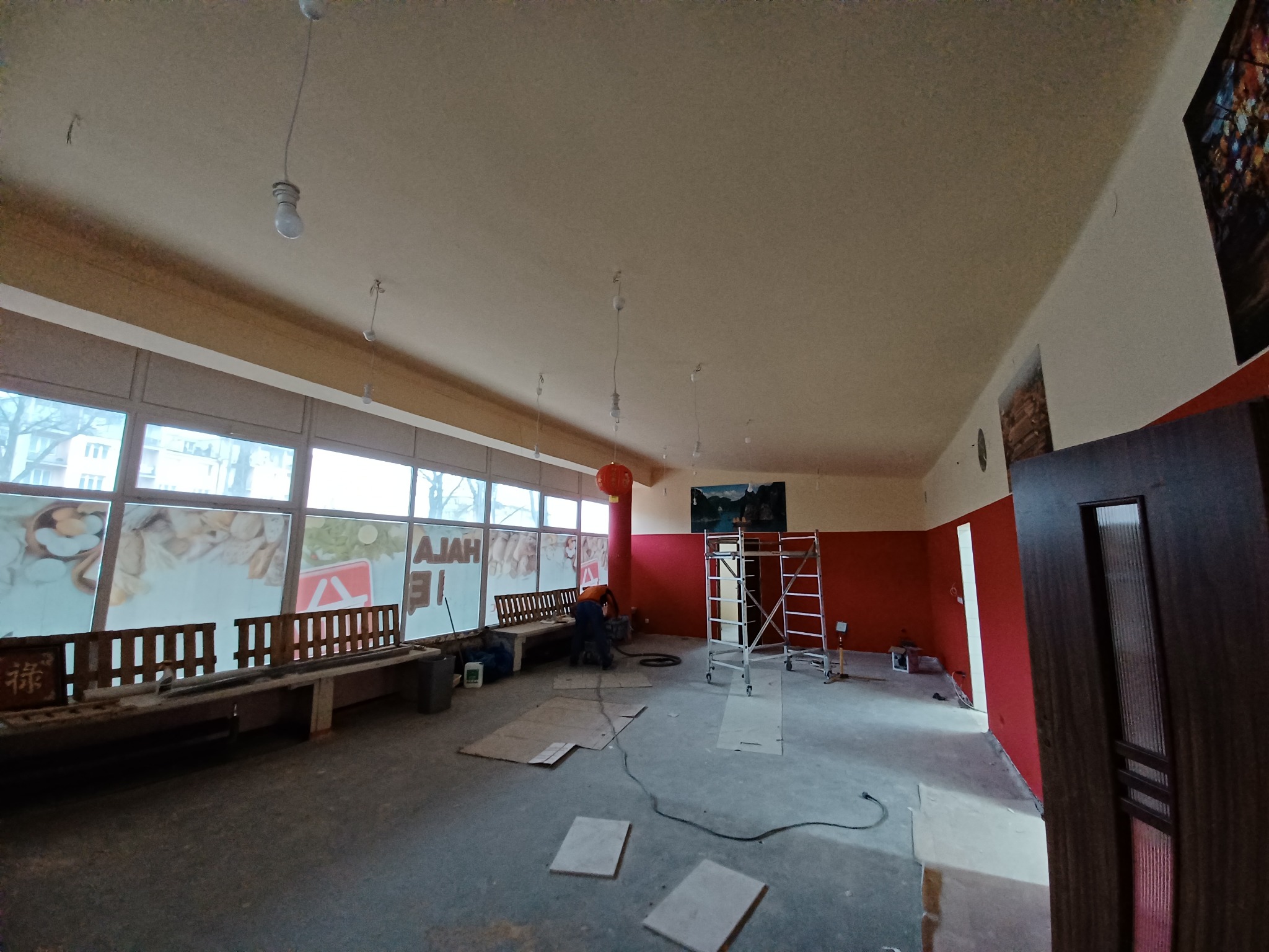 Wnętrze lokalu w trakcie remontu, widoczne rusztowanie, wiszące kable oświetleniowe i pracownik w trakcie pracy, oklejone folią okna, czerwona ściana.