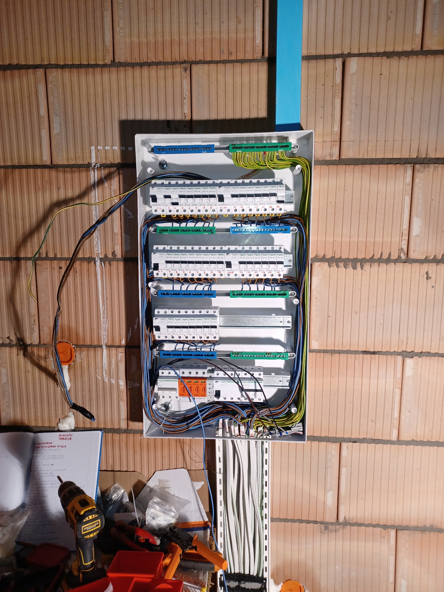 Rozdzielnica elektryczna w trakcie montażu na ścianie z cegły, widoczne przewody, listwa montażowa, narzędzia i notatki na blacie roboczym.