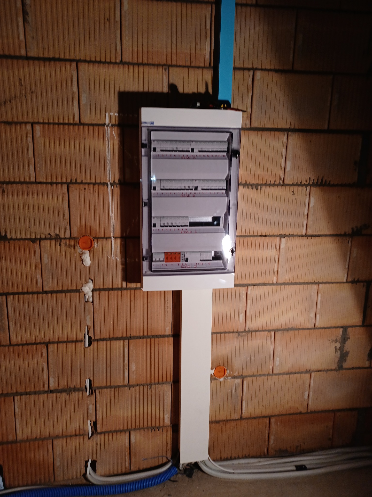 Rozdzielnica elektryczna z zabezpieczeniami zamontowana na ceglanej ścianie, widoczne przewody w peszlach i puszki instalacyjne.