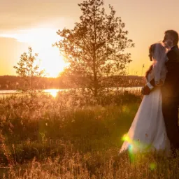Para młoda obejmująca się na tle zachodzącego słońca nad jeziorem, otoczona wysoką trawą i drzewami.