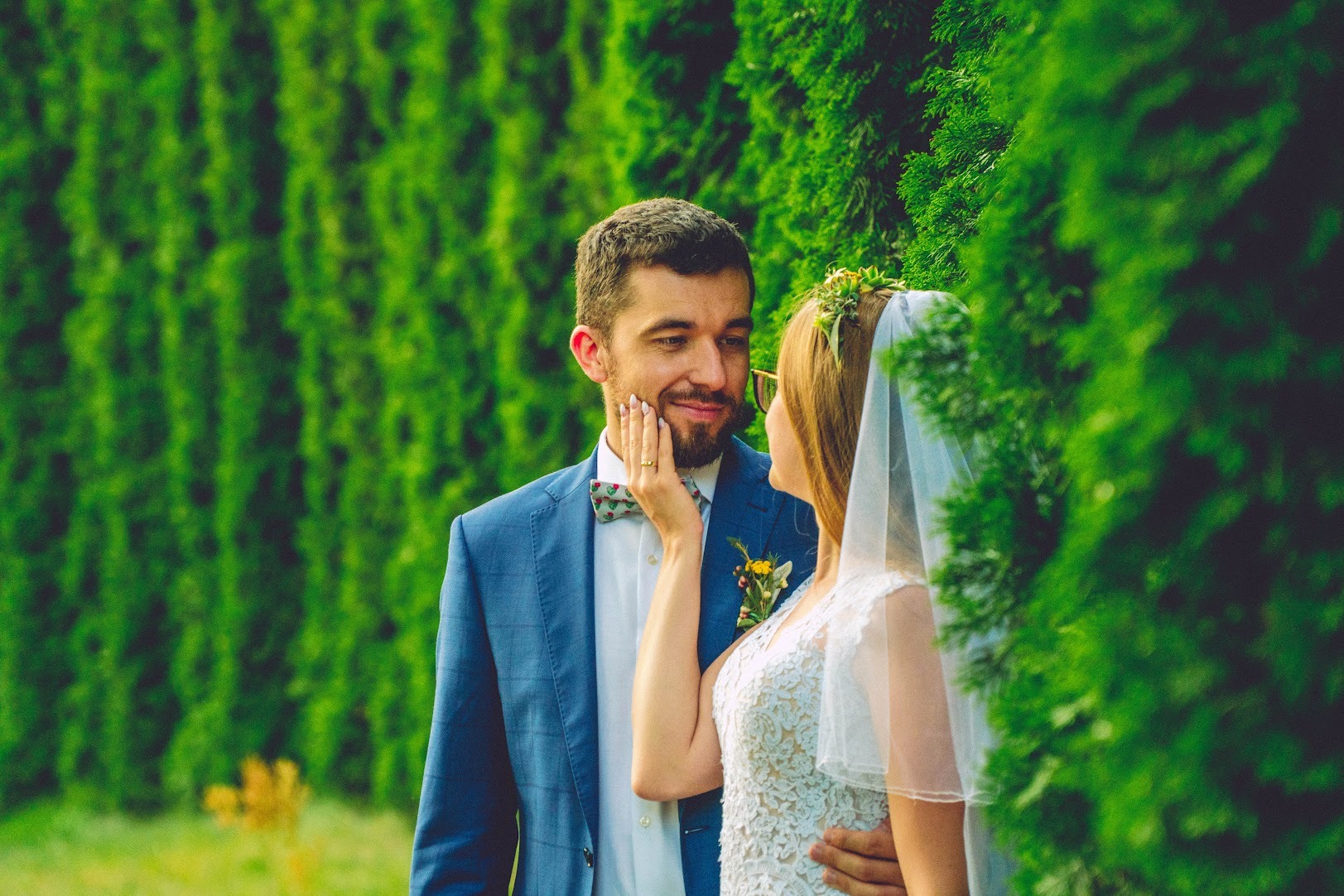Uśmiechnięta para młoda w objęciach na tle zielonej ściany z tui. Panna młoda w koronkowej sukni i wianku z welonem dotyka policzka pana młodego w niebieskiej marynarce i muszce w kwiaty.