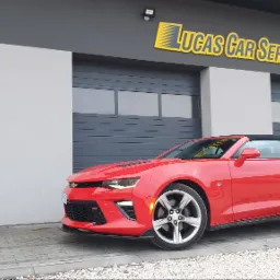 Czerwony kabriolet Chevrolet Camaro zaparkowany przed budynkiem warsztatu samochodowego Lucas Car Service z szarymi panelami i żółtym logo.