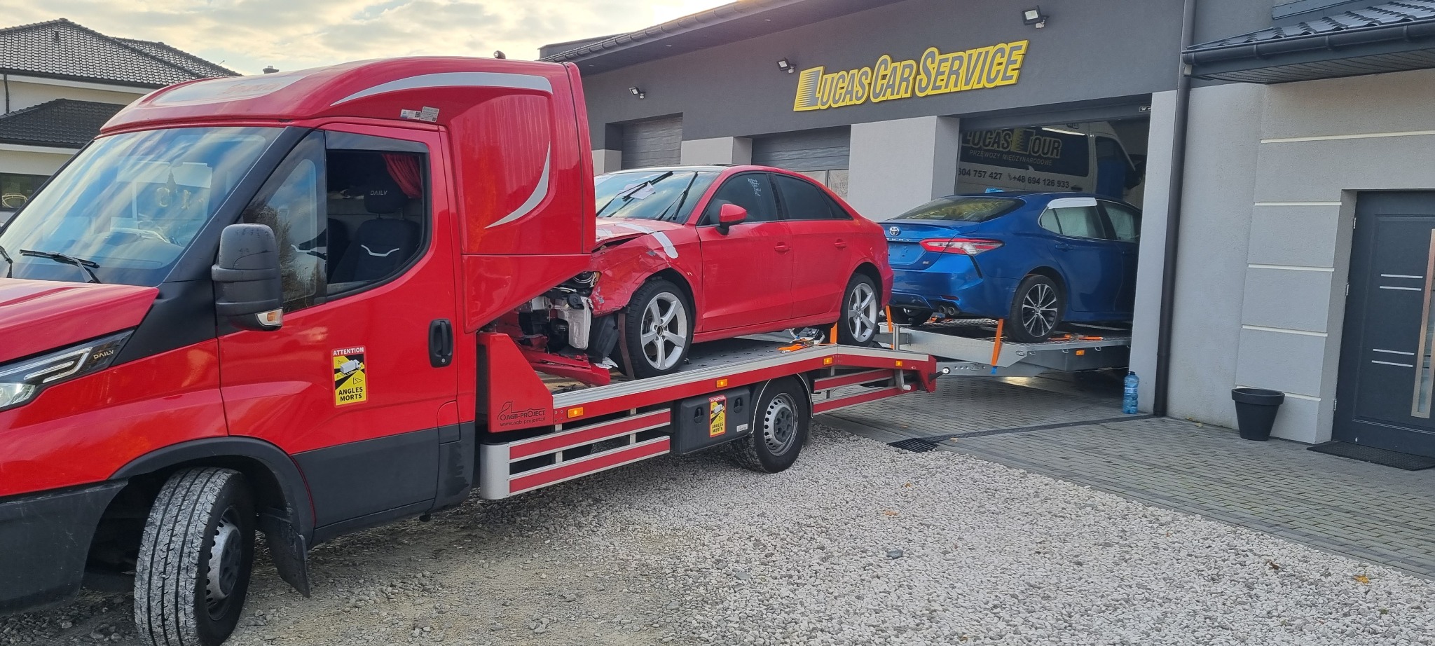 Czerwona laweta z uszkodzonym czerwonym Audi A3 na platformie, zaparkowana przed warsztatem samochodowym Lucas Car Service, w tle niebieska Toyota na drugiej lawecie.
