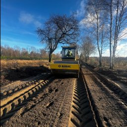 DAKON prace ziemne i usługi komunalno transportowe - Żółty walec drogowy Bomag ubija ziemię na tle drzew i błękitnego nieba. Widoczne ślady opon. Operator w kabinie. Prace ziemne w toku.