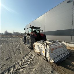 DAKON prace ziemne i usługi komunalno transportowe - Czerwony traktor z białym urządzeniem do wyrównywania terenu, widok z tyłu, na tle szarego budynku magazynowego i błękitnego nieba. Wyrównany teren z lewej strony.
