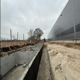 DAKON prace ziemne i usługi komunalno transportowe - Betonowe segmenty w ziemi, prawdopodobnie zbiornik retencyjny, z widocznym wykopem i budynkiem w tle.