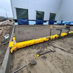 DAKON prace ziemne i usługi komunalno transportowe - Wyrównywanie betonu przy użyciu żółtej listwy wibracyjnej GEPLER na placu budowy, z widocznymi łopatami i fragmentem budynku magazynowego w tle.