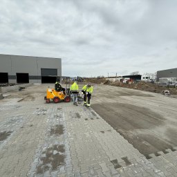 DAKON prace ziemne i usługi komunalno transportowe - Układanie szarej kostki brukowej na dużej powierzchni przemysłowej z użyciem specjalistycznej maszyny i ekipy w odblaskowych kamizelkach. W tle hala magazynowa.