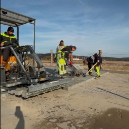DAKON prace ziemne i usługi komunalno transportowe - Ekipa brukarska układa kostkę za pomocą maszyny na placu budowy; widoczni pracownicy w odblaskowych ubraniach i częściowo ułożona nawierzchnia.