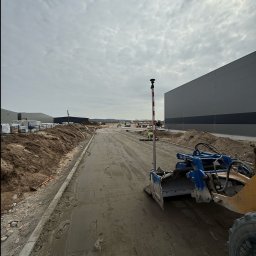 DAKON prace ziemne i usługi komunalno transportowe - Niwelacja terenu pod budowę hali. W tle hałdy ziemi i budynki. Na pierwszym planie maszyna do niwelacji i tyczka geodezyjna. Pochmurny dzień.