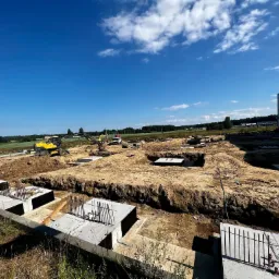 Fundamenty pod budynek przemysłowy w trakcie budowy, widoczne wykopy, betonowe ławy fundamentowe ze zbrojeniami i maszyny budowlane w tle. Słoneczny dzień.