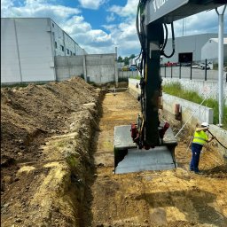 DAKON prace ziemne i usługi komunalno transportowe - Praca ziemne: koparka Volvo w trakcie wykonywania wykopu liniowego. Pracownik w kasku i kamizelce odblaskowej wyrównuje skarpę. W tle budynek przemysłowy i błękitne niebo.