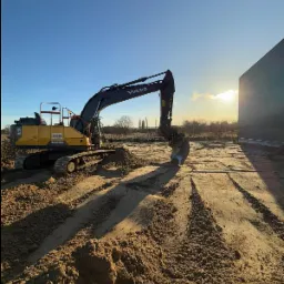 Żółta koparka Volvo na placu budowy przy budynku, w tle zachodzące słońce i drzewa. Widoczne ślady opon na piasku.