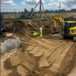 DAKON prace ziemne i usługi komunalno transportowe - Żółta koparka i zagęszczarka w wykopie z rurami, pracownik w odblaskowej kamizelce, teren przygotowany pod dalsze prace ziemne.