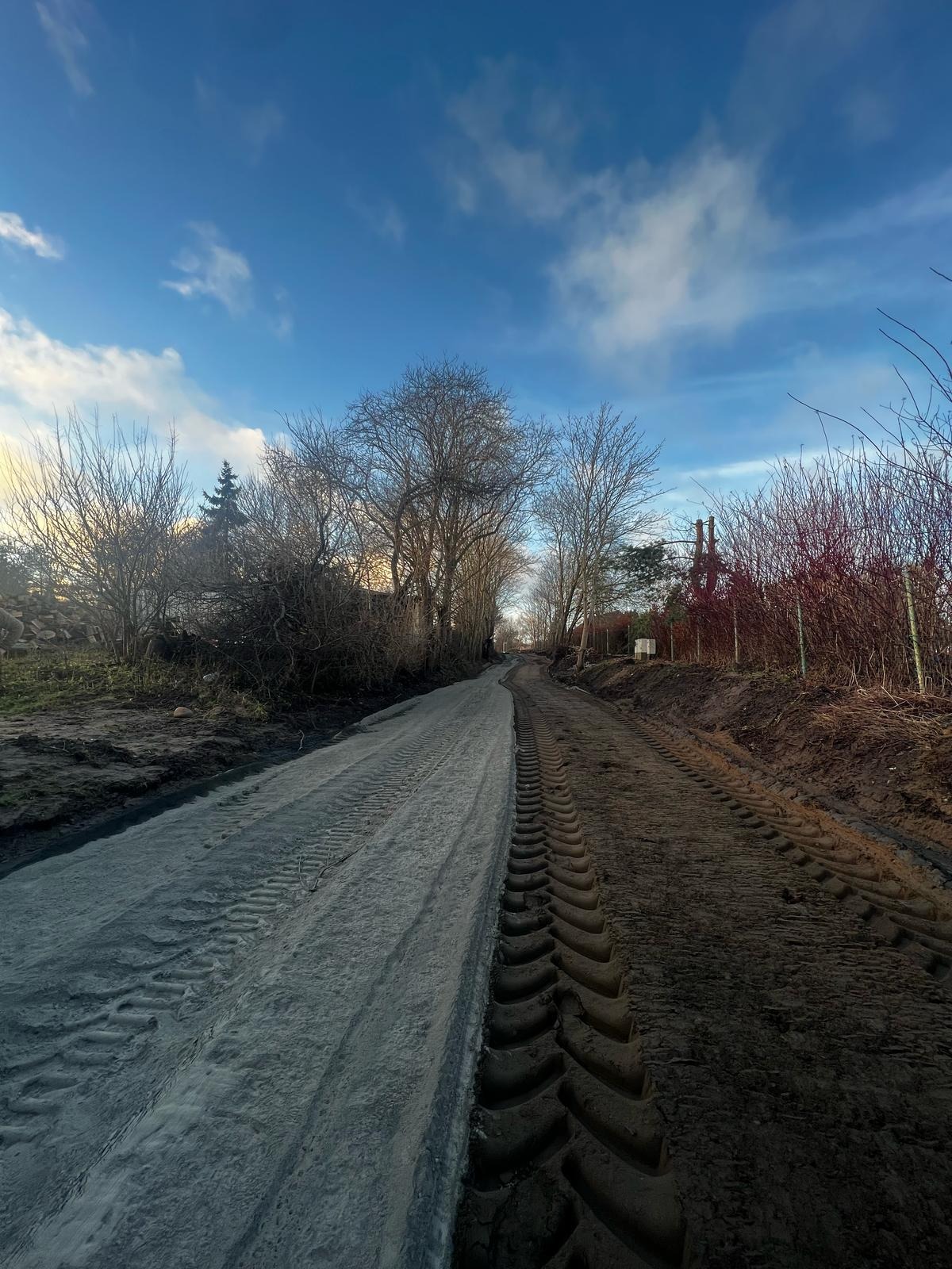 Połowa drogi utwardzona, widoczne ślady opon, druga połowa z ziemią, błękitne niebo z chmurami w tle. Ścieżka prowadzi w dal.