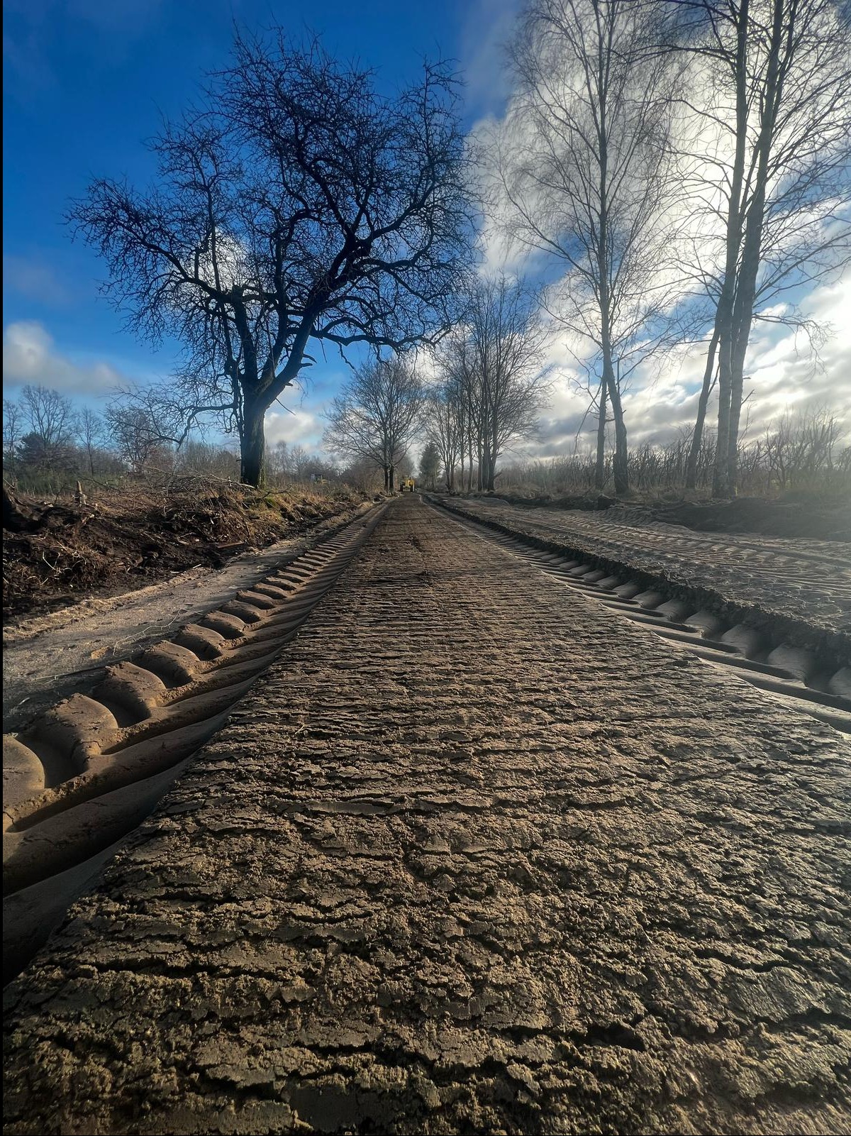 Wyrównany teren z wyraźnymi śladami po maszynie budowlanej, drzewa w tle pod błękitnym niebem z chmurami. Widok w perspektywie.