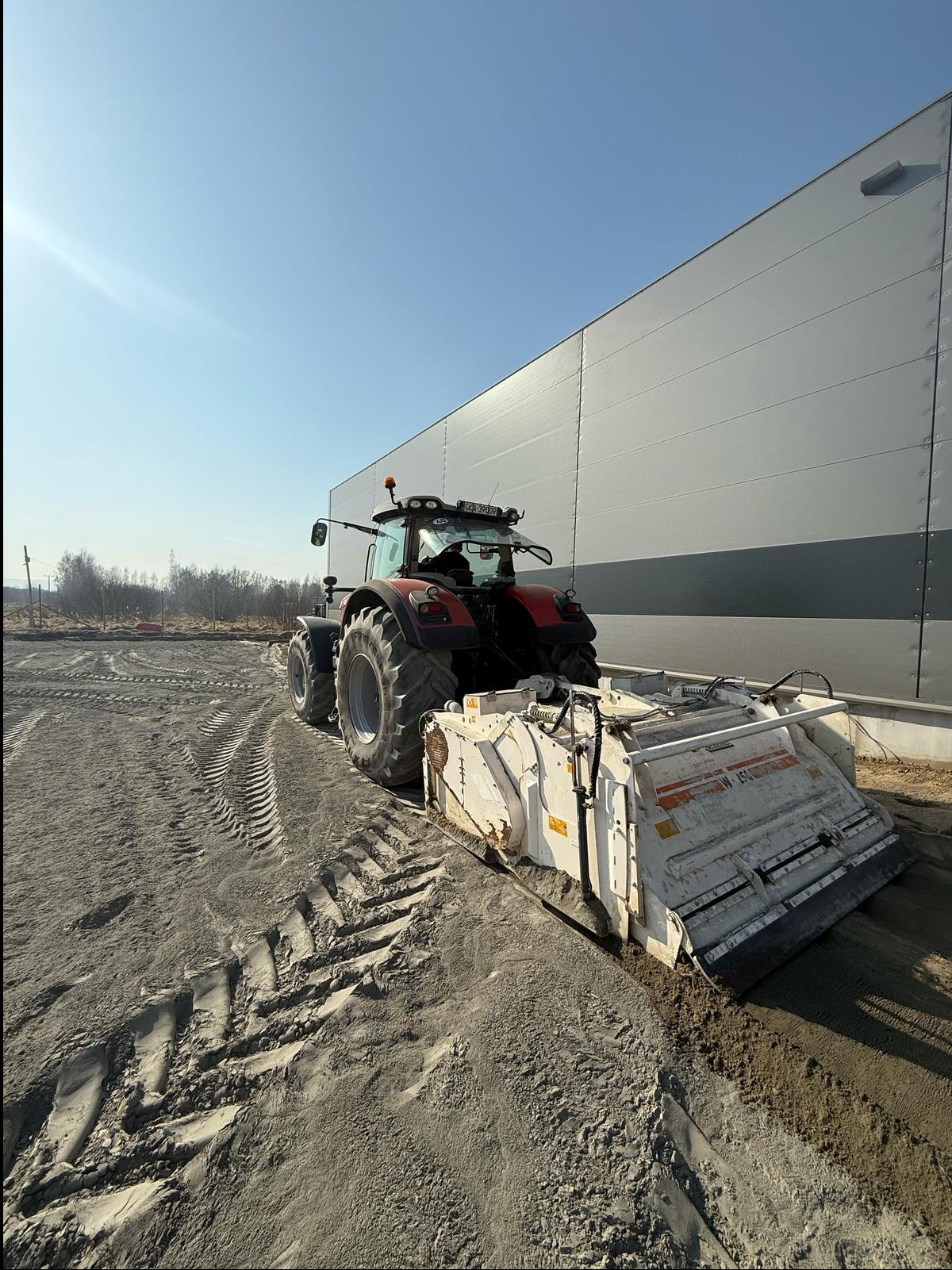 Czerwony traktor z białym urządzeniem do wyrównywania terenu, widok z tyłu, na tle szarego budynku magazynowego i błękitnego nieba. Wyrównany teren z lewej strony.