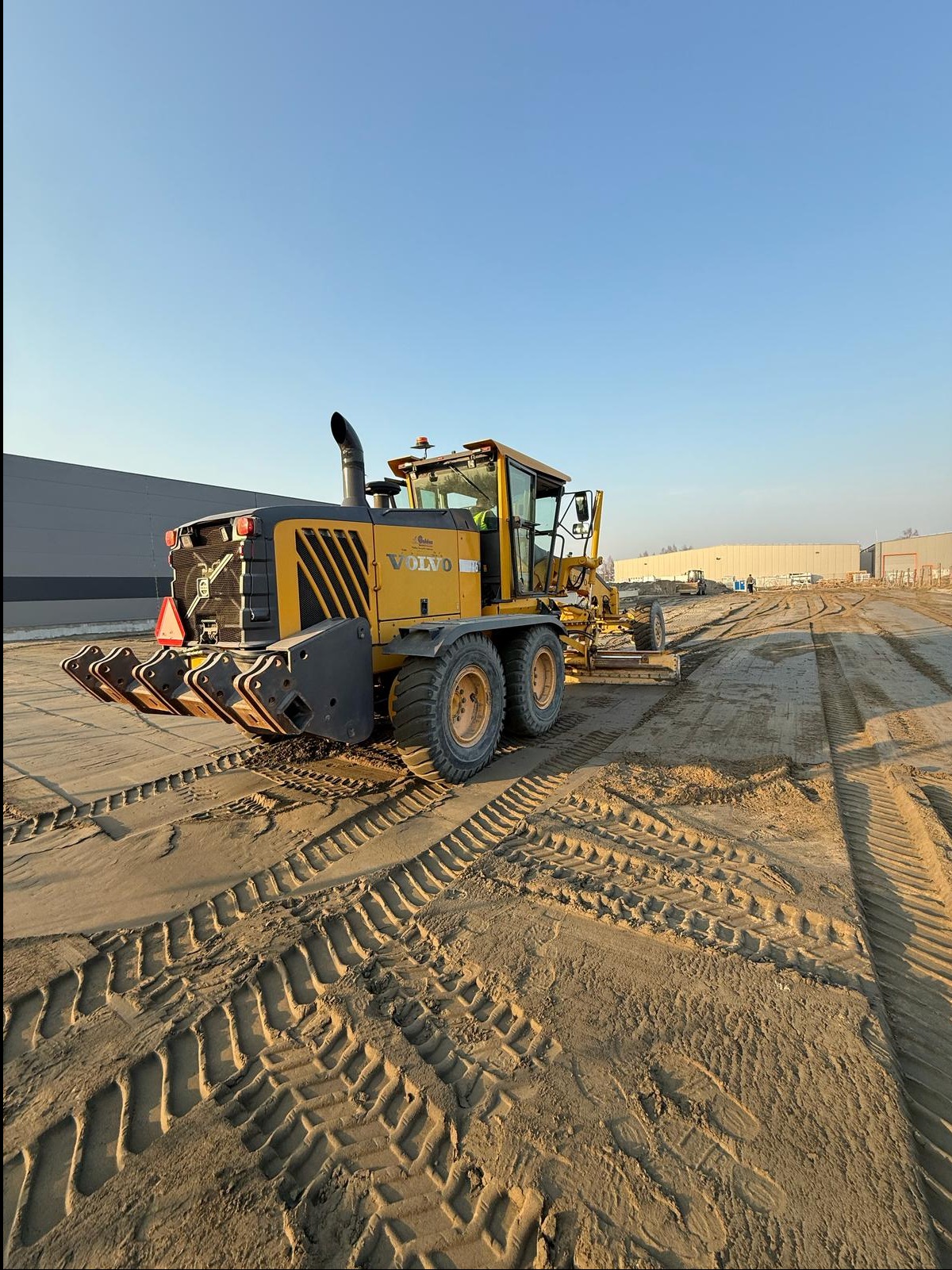 Żółty równiarka VOLVO na piaszczystym terenie budowy, widoczne ślady opon, budynki w tle. Maszyna gotowa do pracy.