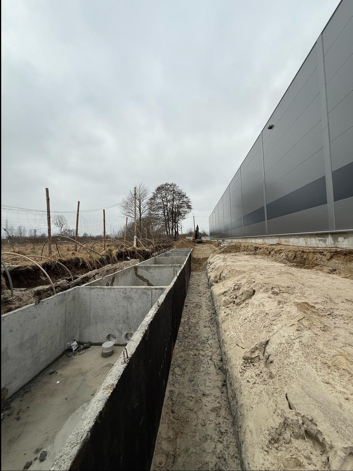 Betonowe segmenty w ziemi, prawdopodobnie zbiornik retencyjny, z widocznym wykopem i budynkiem w tle.