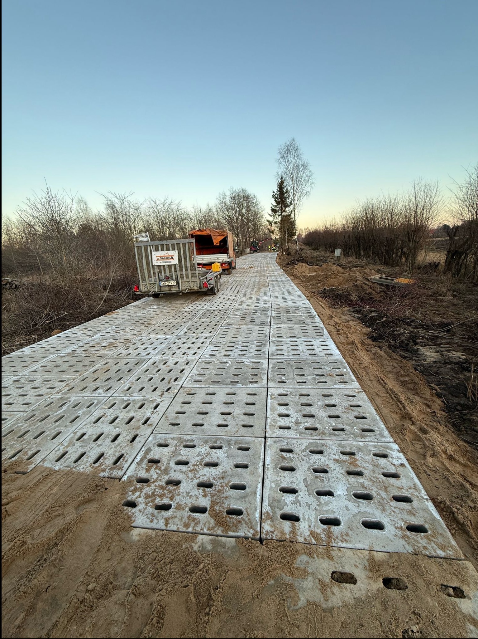 Utwardzona droga z betonowych płyt ażurowych, widoczny przyczepiony wózek i ciężarówka w oddali, brązowe zarośla po bokach drogi, niebo w odcieniach błękitu i różu.