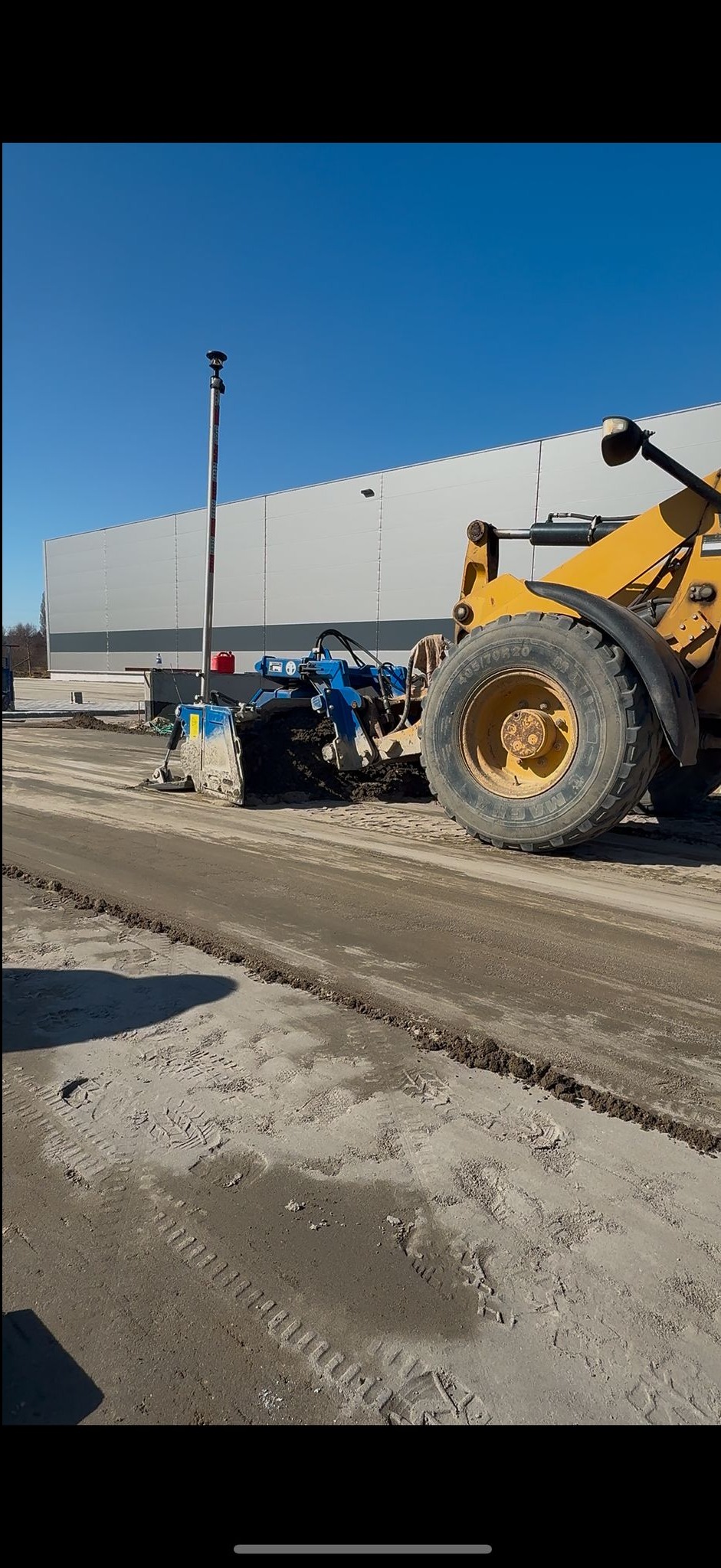 Żółta równiarka z niebieskim modułem niwelacyjnym wyrównuje teren pod budowę. W tle hala magazynowa i błękitne niebo. Widoczne ślady opon na piasku.