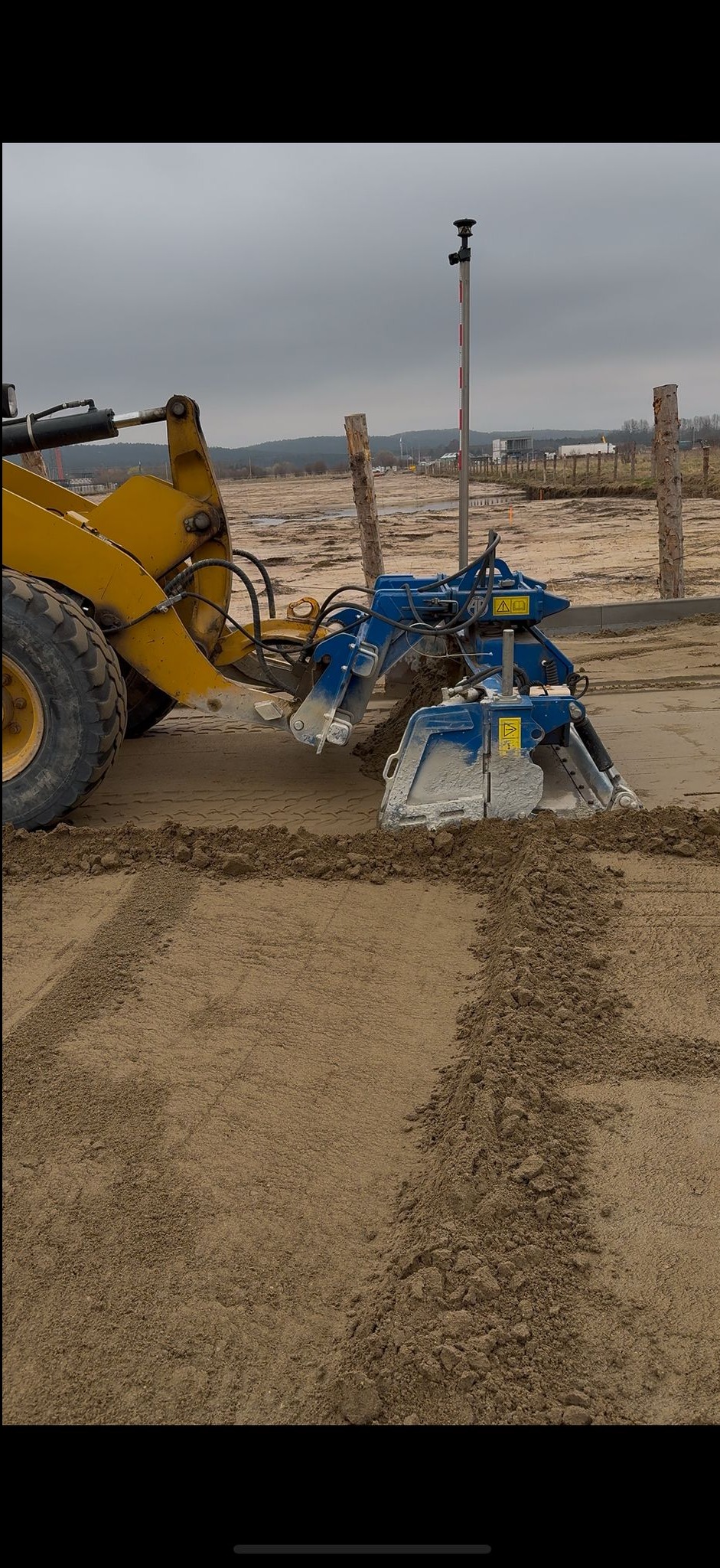 Żółta ładowarka z niebieskim osprzętem profiluje teren piaskowy, tworząc równe linie. W tle widoczny teren z drewnianymi słupkami i budynkami.