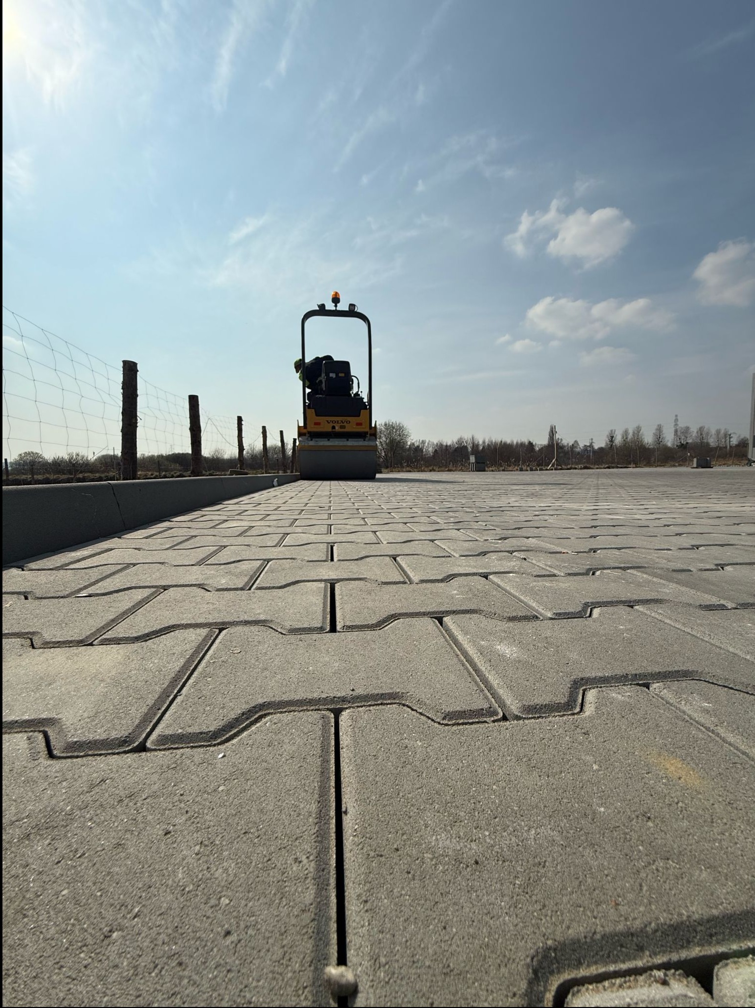 Maszyna do ubijania kostki brukowej na nowo ułożonej nawierzchni z szarej kostki, widok z dołu. W tle siatka ogrodzeniowa i błękitne niebo.