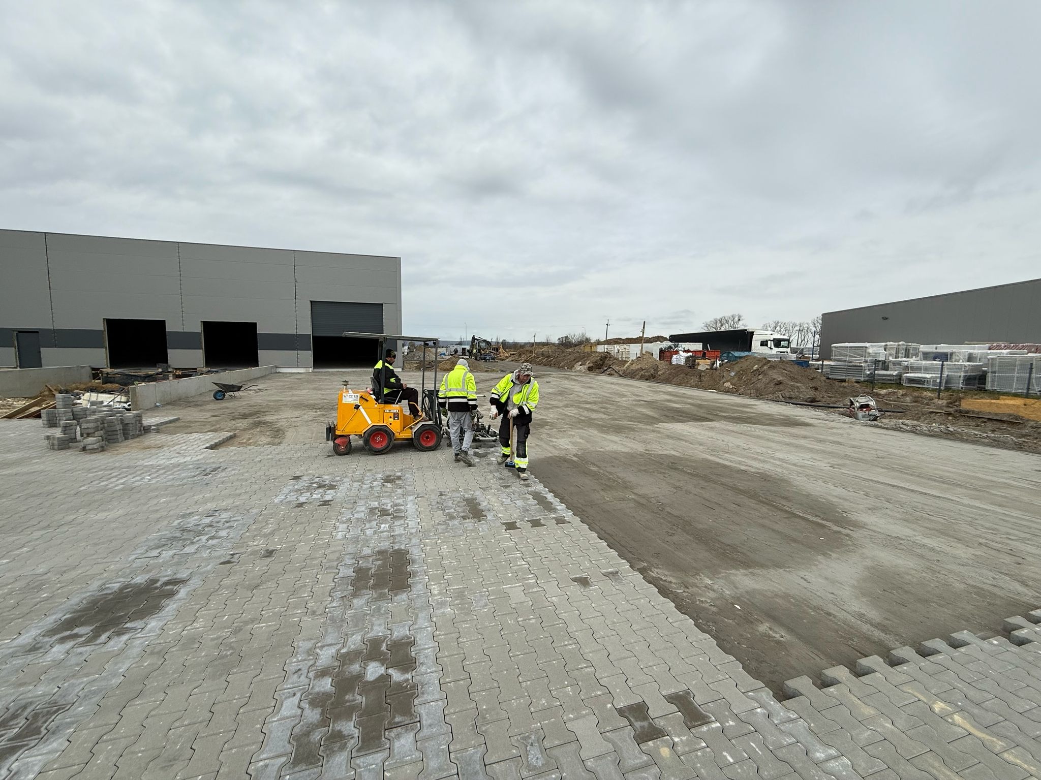 Układanie szarej kostki brukowej na dużej powierzchni przemysłowej z użyciem specjalistycznej maszyny i ekipy w odblaskowych kamizelkach. W tle hala magazynowa.