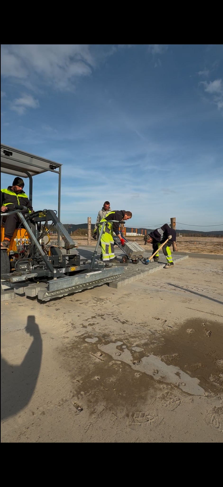 Ekipa brukarska układa kostkę za pomocą maszyny na placu budowy; widoczni pracownicy w odblaskowych ubraniach i częściowo ułożona nawierzchnia.