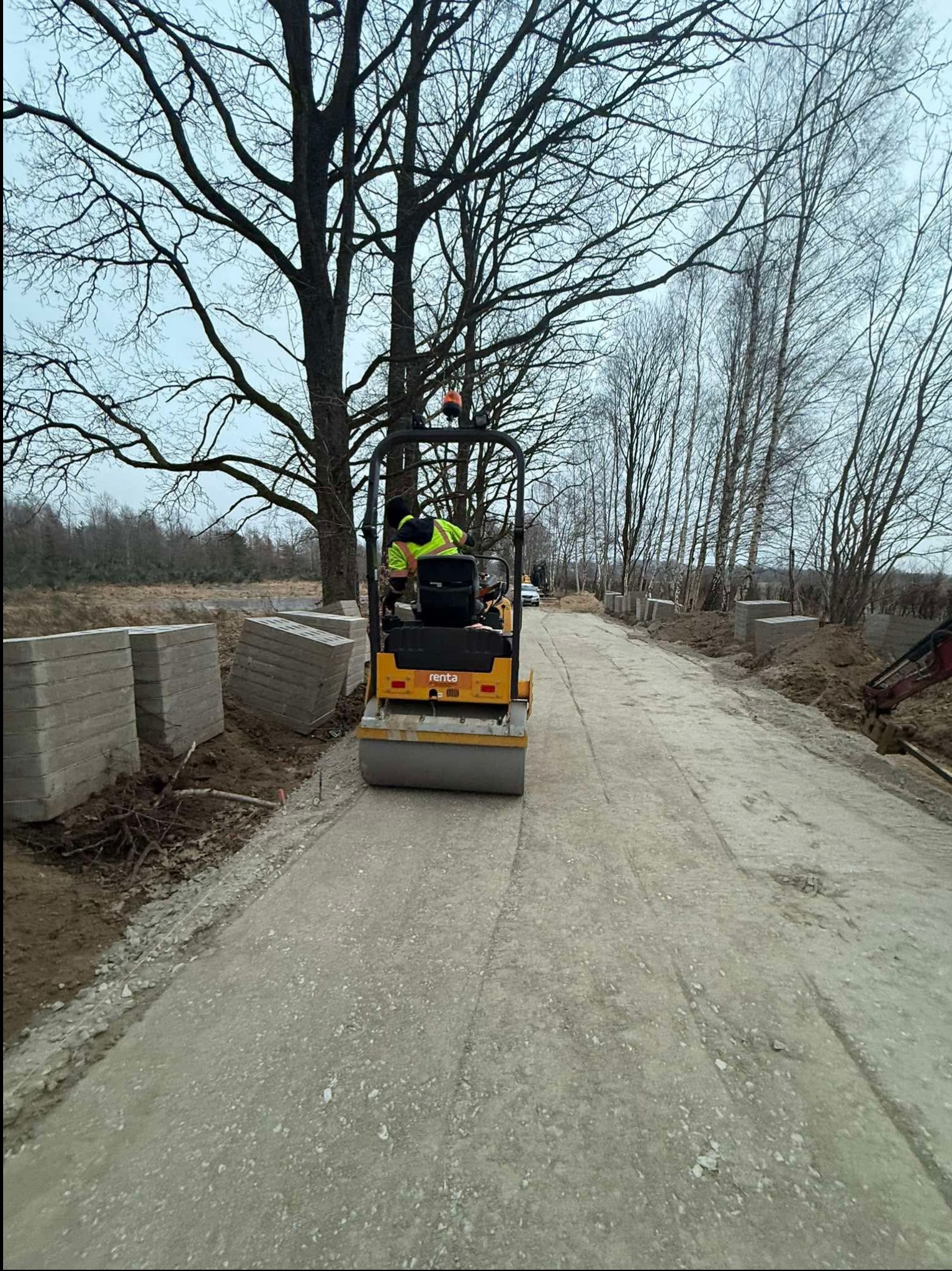 Walec drogowy ubija szutrową nawierzchnię. Po bokach ułożone betonowe bloczki, drzewa bez liści w tle. Praca w toku, operator w odblaskowej kamizelce.