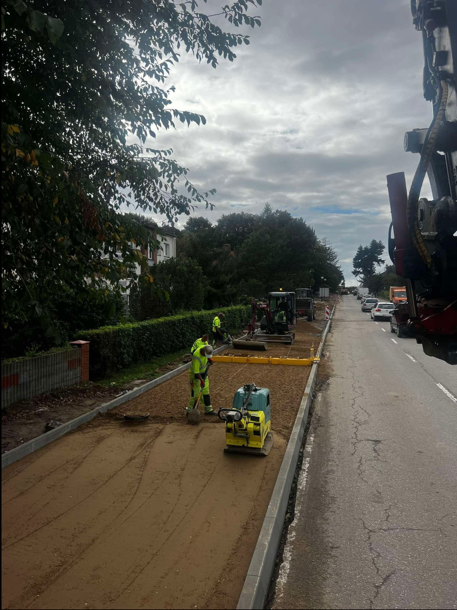 Prace drogowe w toku: robotnicy w odblaskowych ubraniach wyrównują piasek ubijarką, w tle maszyny budowlane i ruch uliczny. Dzień pochmurny, krajobraz miejski.