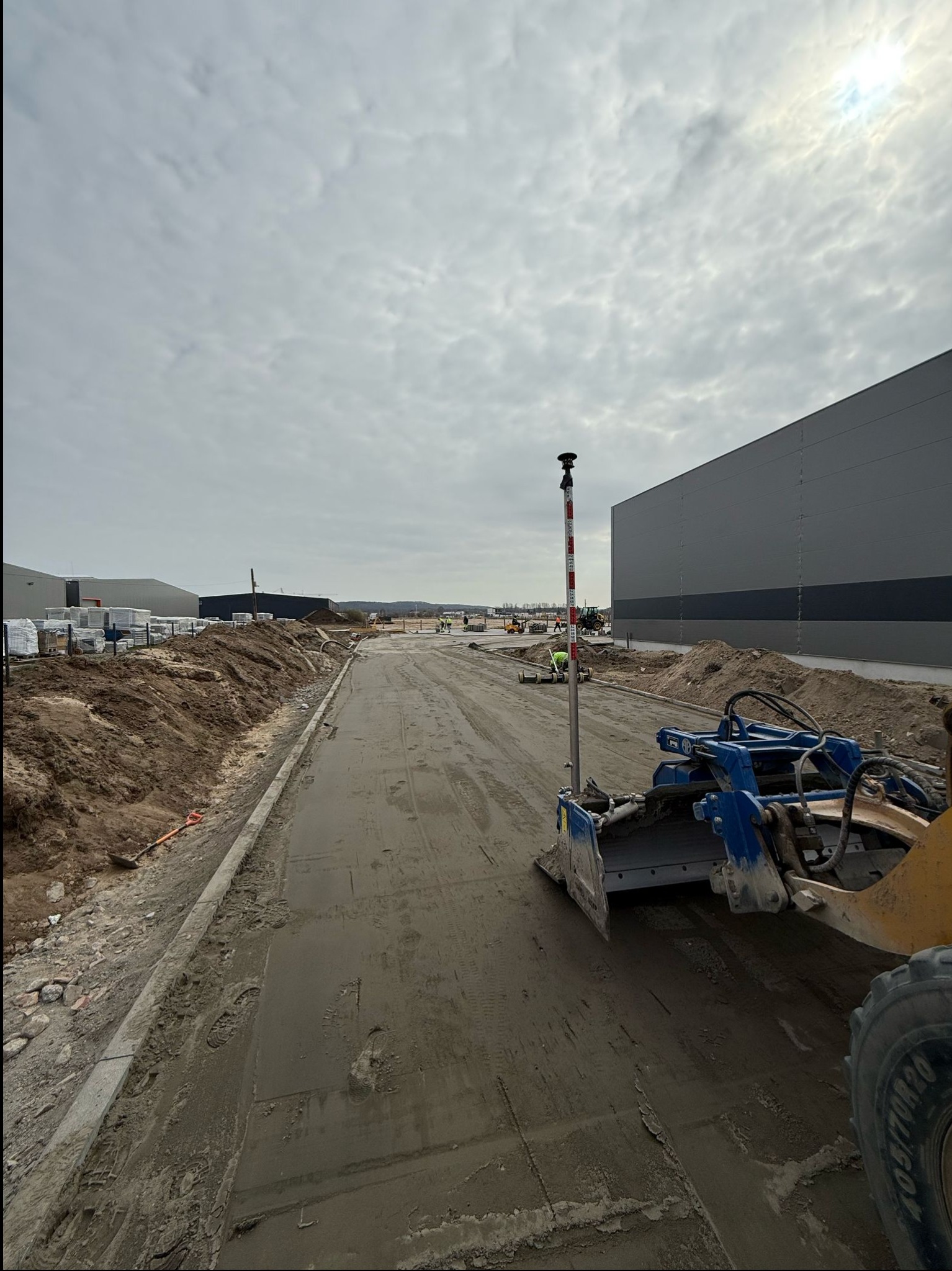 Niwelacja terenu pod budowę hali. W tle hałdy ziemi i budynki. Na pierwszym planie maszyna do niwelacji i tyczka geodezyjna. Pochmurny dzień.