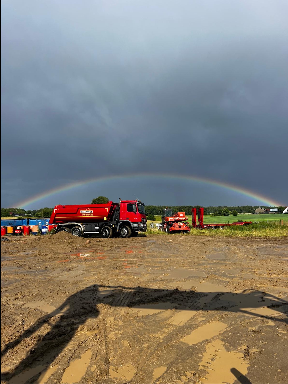 Czerwona wywrotka na błotnistym terenie budowy pod tęczą, z widocznym sprzętem budowlanym w tle i ciemnym, burzowym niebem.
