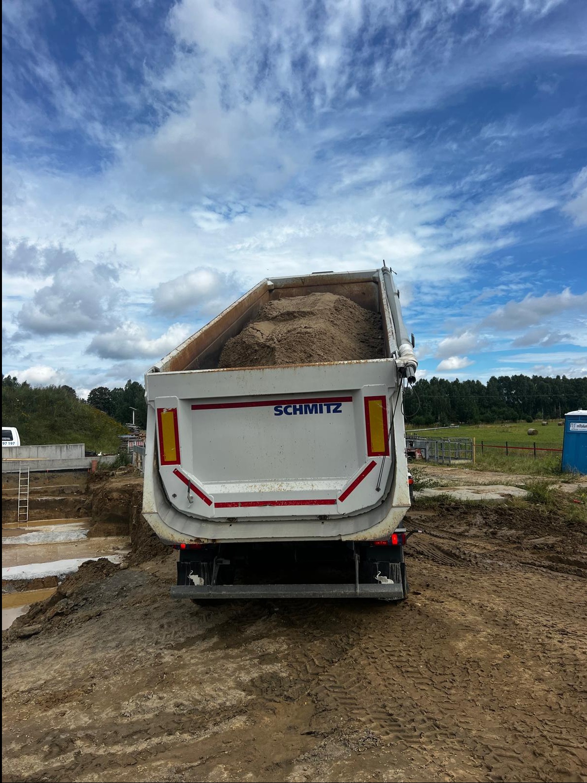Ciężarówka Schmitz z piaskiem na placu budowy, widok z tyłu, w tle wykop pod fundamenty i błękitne niebo z chmurami.