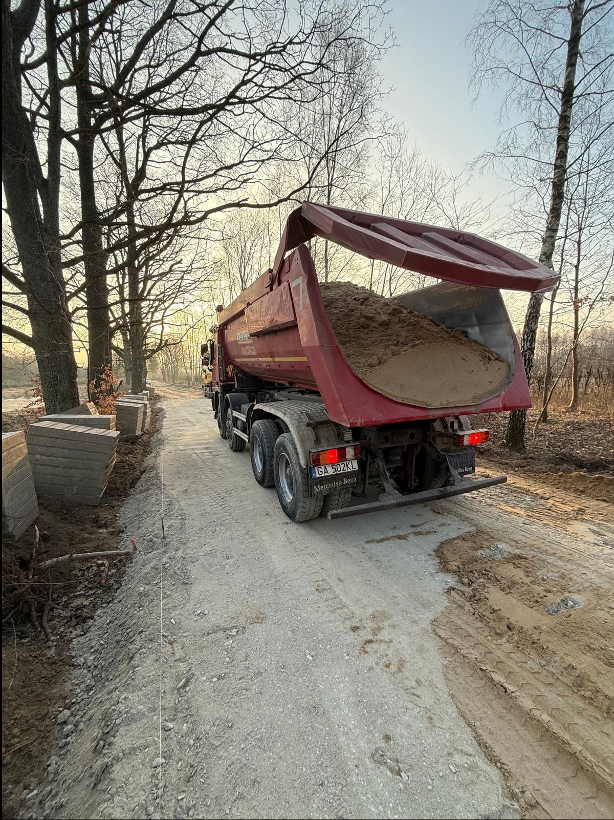 Ciężarówka wywrotka z otwartą skrzynią, wypełniona piaskiem, stoi na drodze gruntowej obok ułożonych kostek. Prace ziemne w toku, widok z tyłu.