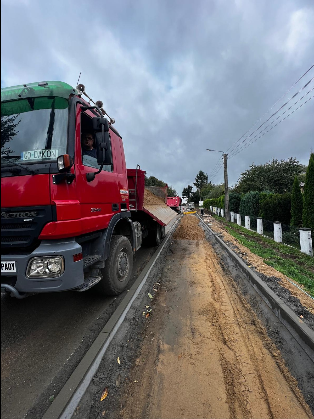 Ciężarówka wyładowuje piasek na przygotowany teren pod budowę drogi w Wejherowie. Widoczne krawężniki i inne maszyny w oddali. Pochmurne niebo.