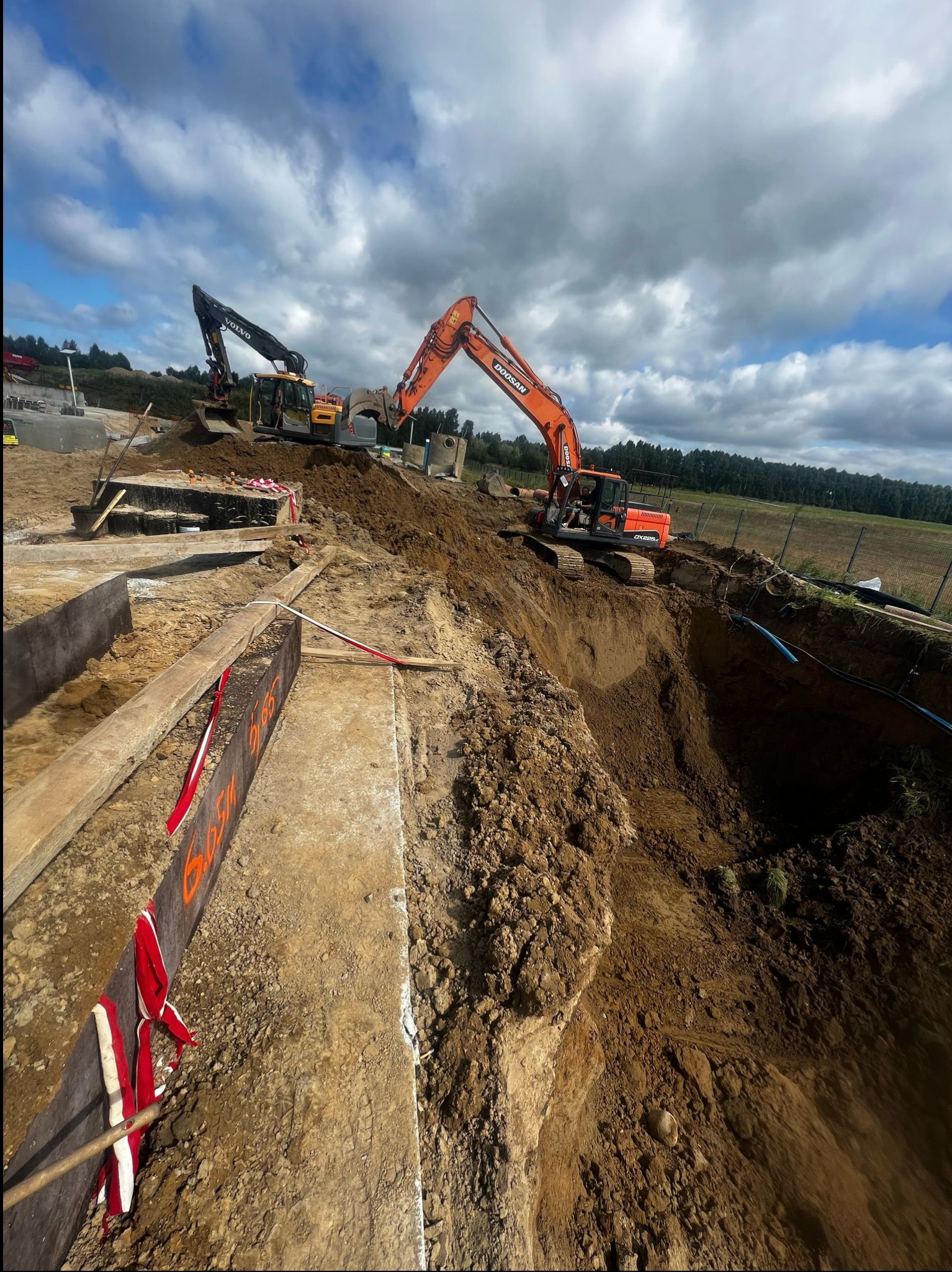 Pomarańczowa koparka Doosan i czarna Volvo pracują na budowie. Widoczne wykopy, szalunki z oznaczeniami i częściowo odsłonięte rury. W tle zielone pola i błękitne niebo.