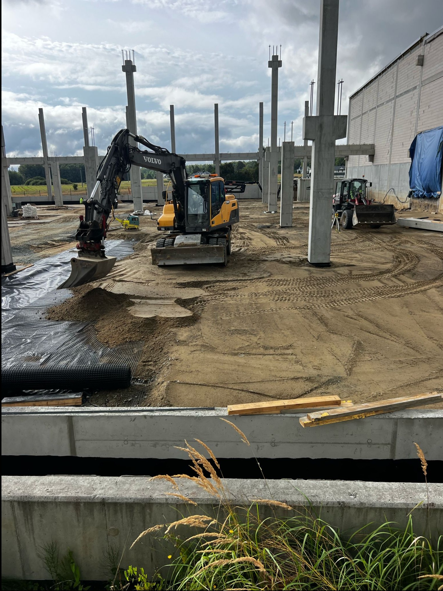 Żółta koparka Volvo na terenie budowy hali z betonowymi kolumnami. Widoczne ślady opon na piasku i czarna siatka. W tle zielony krajobraz pod pochmurnym niebem.